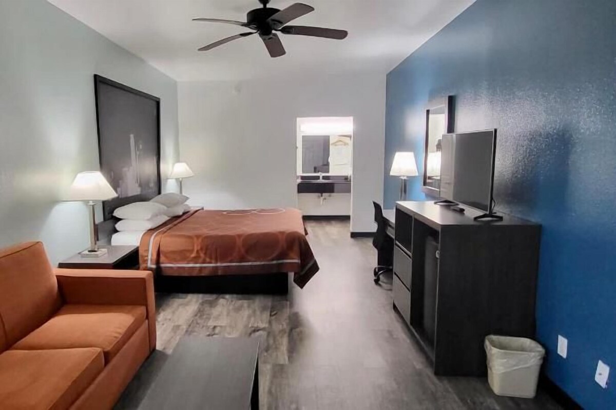 Een hotelkamer met een bed, een bank, een bureau, een televisie en een plafondventilator.
