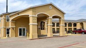 A building with a beige facade, numbered 103, featuring a central entrance with arches, a parking lot, and a red vehicle parked nearby. - Scottish Inns & Suites Forney/East Dallas (Forney)