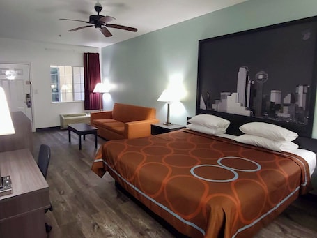 A hotel room with a bed, a desk, a chair, a ceiling fan, and a cityscape painting on the wall.. Scottish Inns & Suites Forney/East Dallas