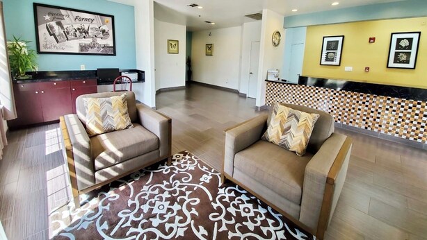 A reception area with a blue wall, a framed welcome sign, a reception desk, and two sofas with patterned cushions. - Scottish Inns & Suites Forney/East Dallas (Forney)