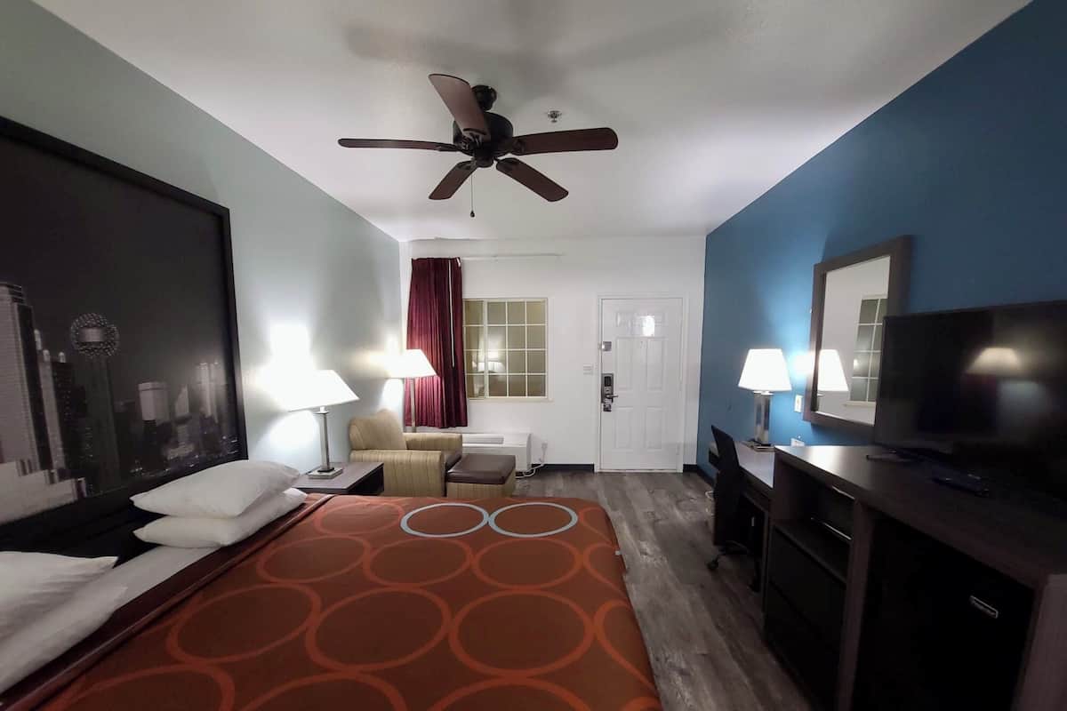 A hotel room with a bed, a desk, a television, a ceiling fan, and a window with a cityscape view.