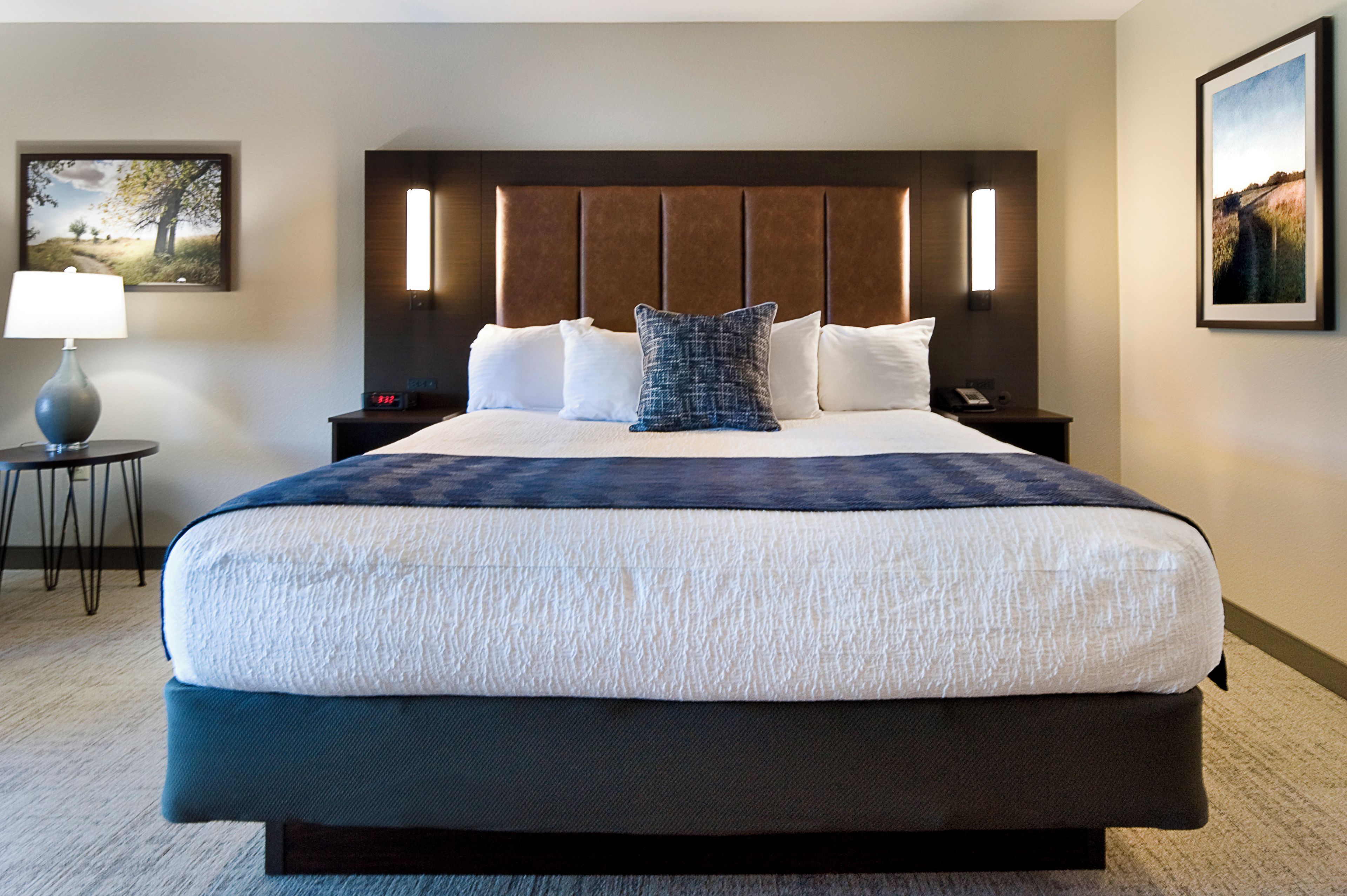 Una habitación de hotel con una cama grande, una mesita de noche con una lámpara, un mueble para el teléfono y dos cuadros en las paredes.