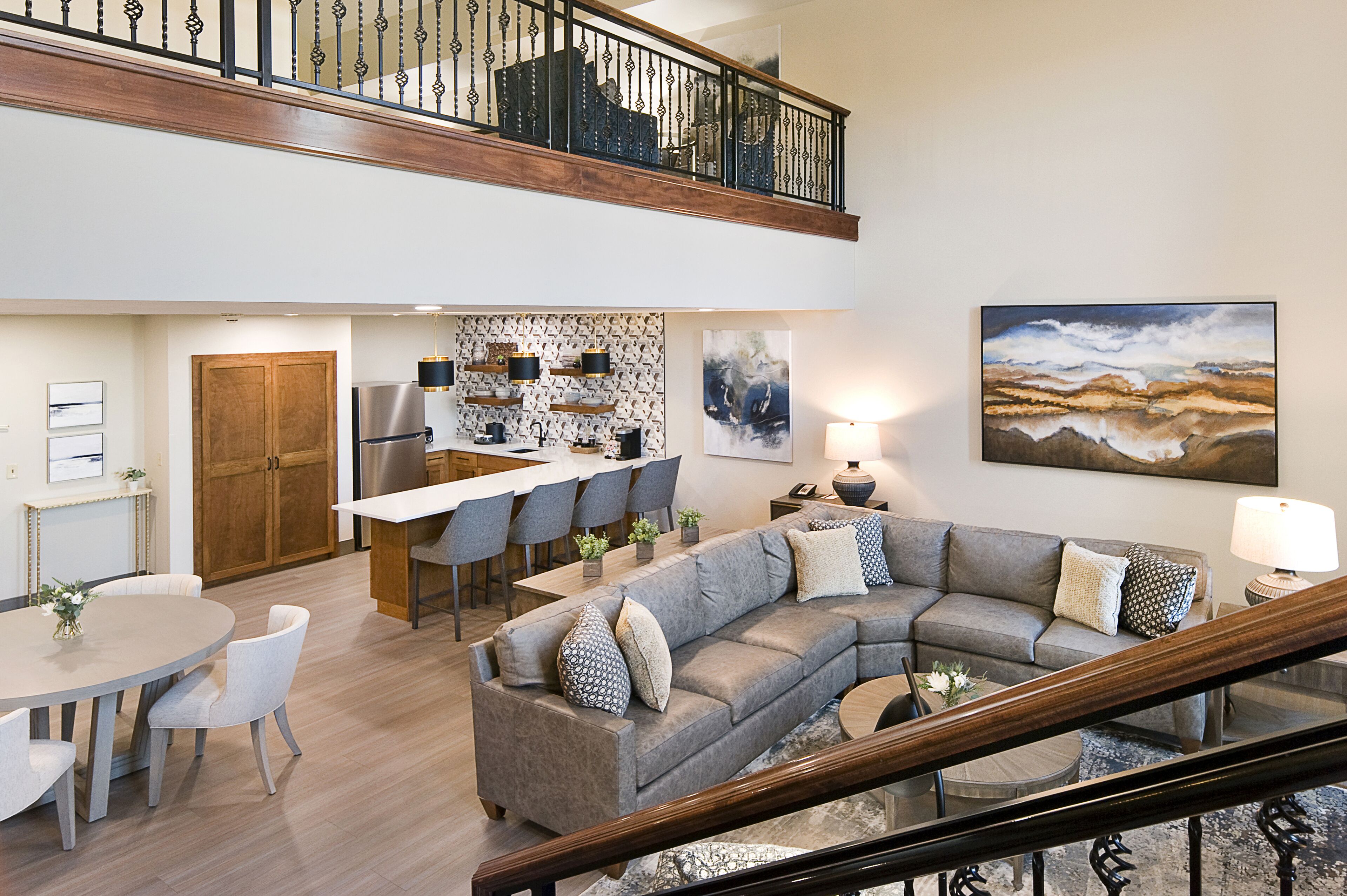 A spacious living area with a large gray sectional sofa, a dining table with chairs, a kitchen area with a bar, and a staircase leading to an upper level.