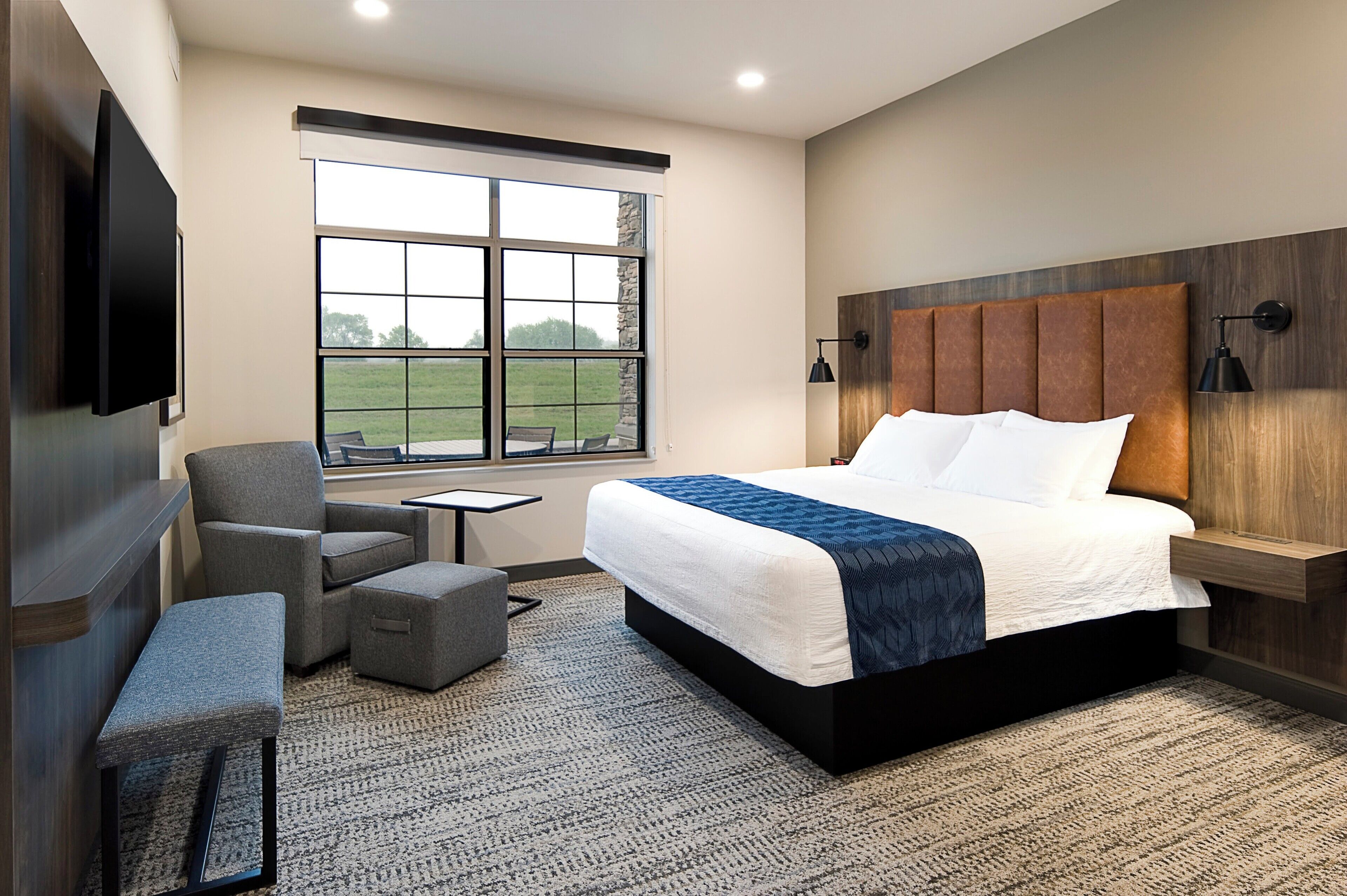A modern hotel room with a large bed, a TV, a chair, a small table, and a window with a view of the outdoors.