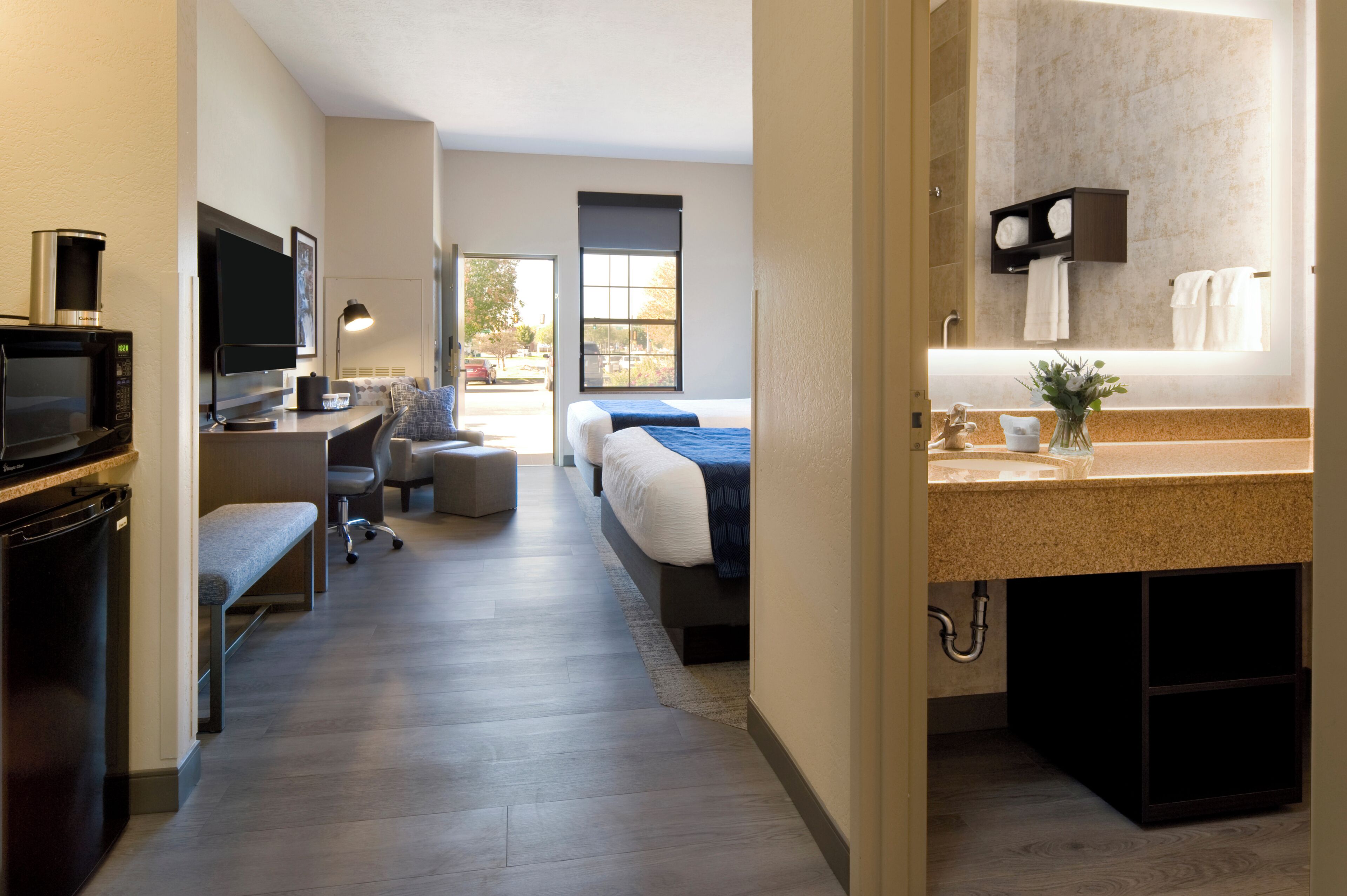 A modern hotel room with a kitchenette, a bed, a desk with a chair, a large window, and a bathroom with a sink and towel rack.
