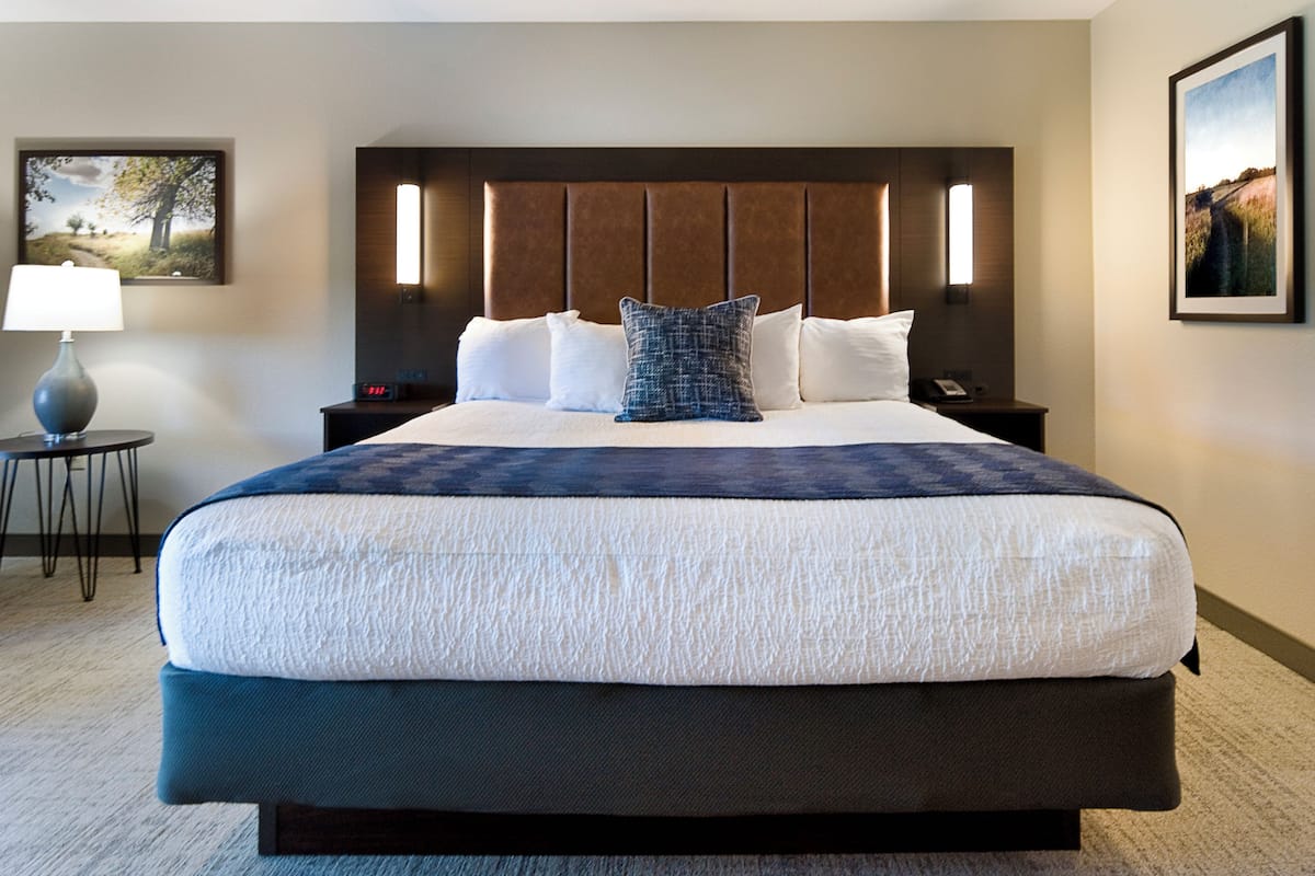 Une chambre d’hôtel avec un grand lit, une table de chevet avec une lampe, une petite table avec un téléphone et deux tableaux encadrés accrochés aux murs.
