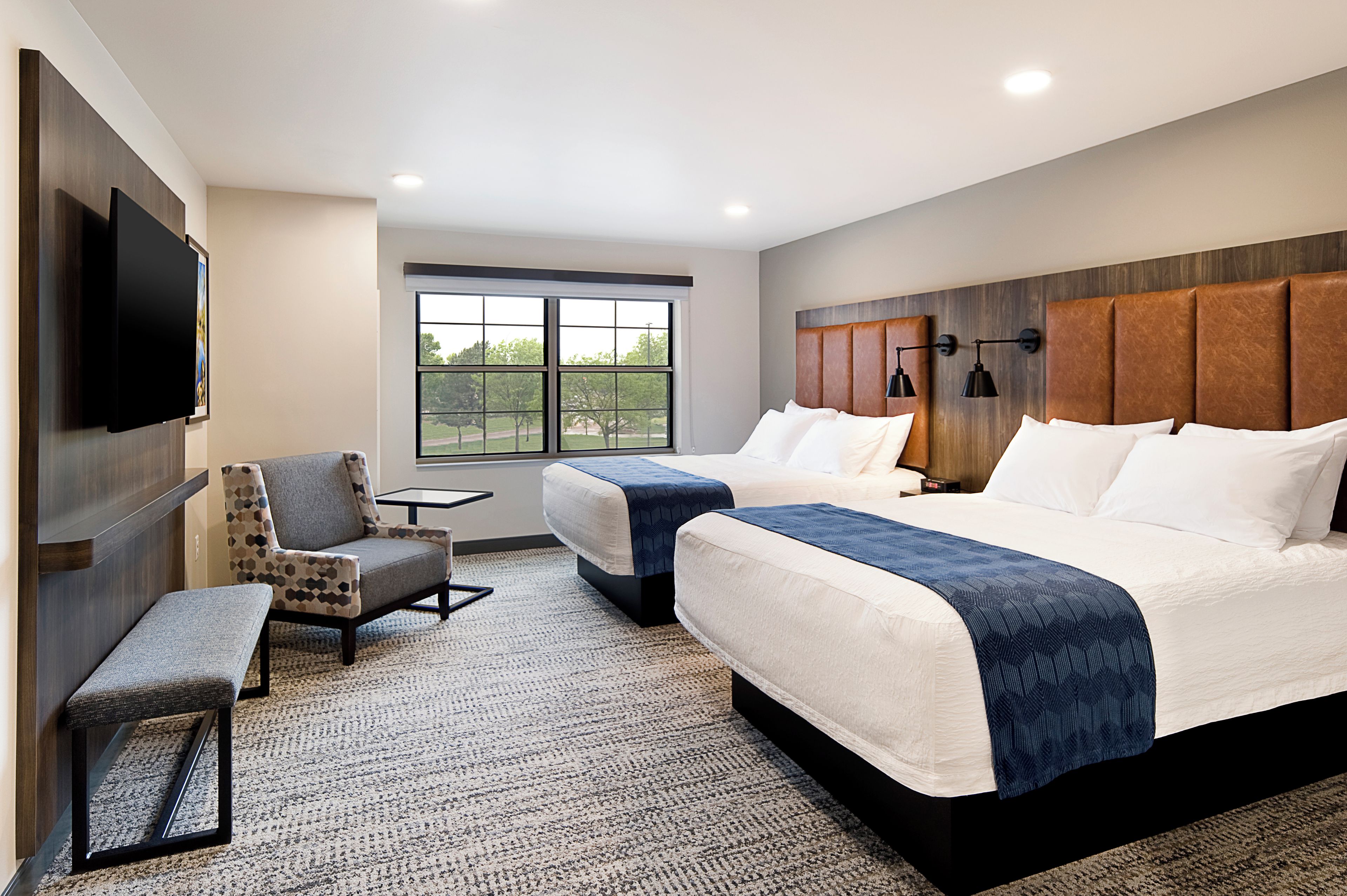 A hotel room with two beds, a TV, a chair, and a window with a view of trees.