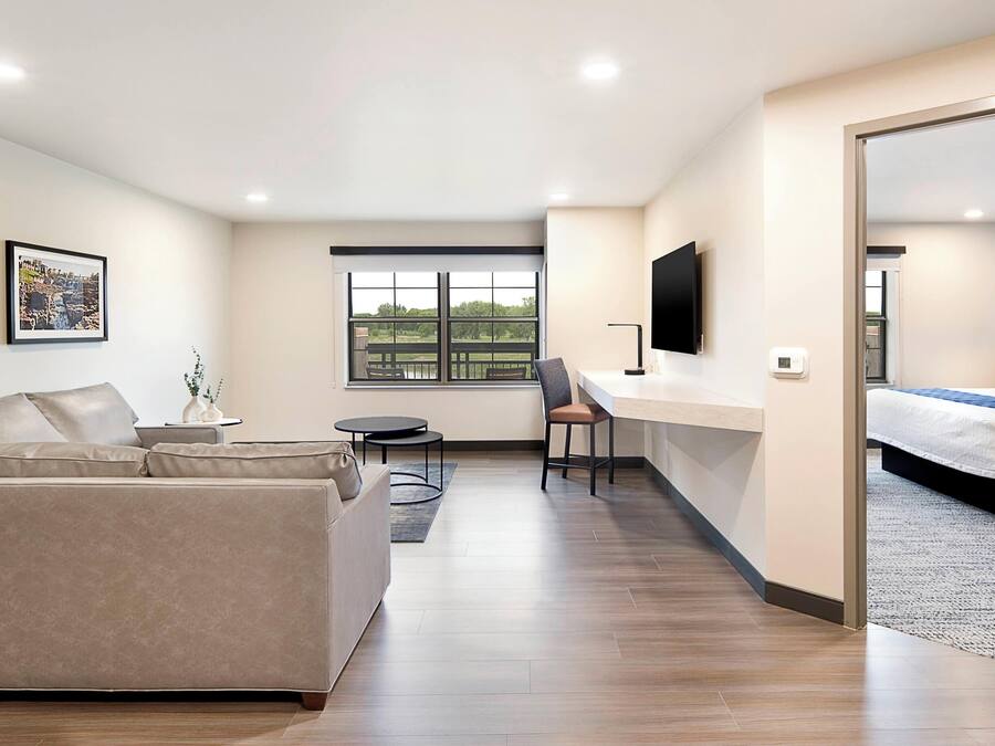 A modern hotel room with a sofa, a desk, a chair, a bed, a TV, and a large window.