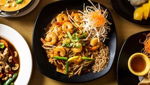 A variety of dishes including a plate of noodles with shrimp, a bowl of soup, and a plate of mango slices.