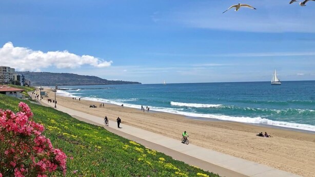 A beach with a paved walkway, people walking and cycling, and a sailboat in the distance.