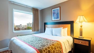 A hotel room with a large bed, a nightstand with a lamp, a window with a view of the city, and a framed picture on the wall.