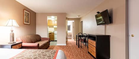 Habitación de hotel con cama, escritorio, silla, televisión y una kitchenette al fondo.
