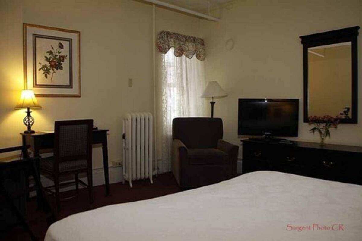 Habitación de hotel con cama, escritorio con silla, televisor, lámpara, radiador y espejo.
