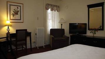 A hotel room with a bed, a desk with a chair, a TV, a lamp, a radiator, and a mirror.