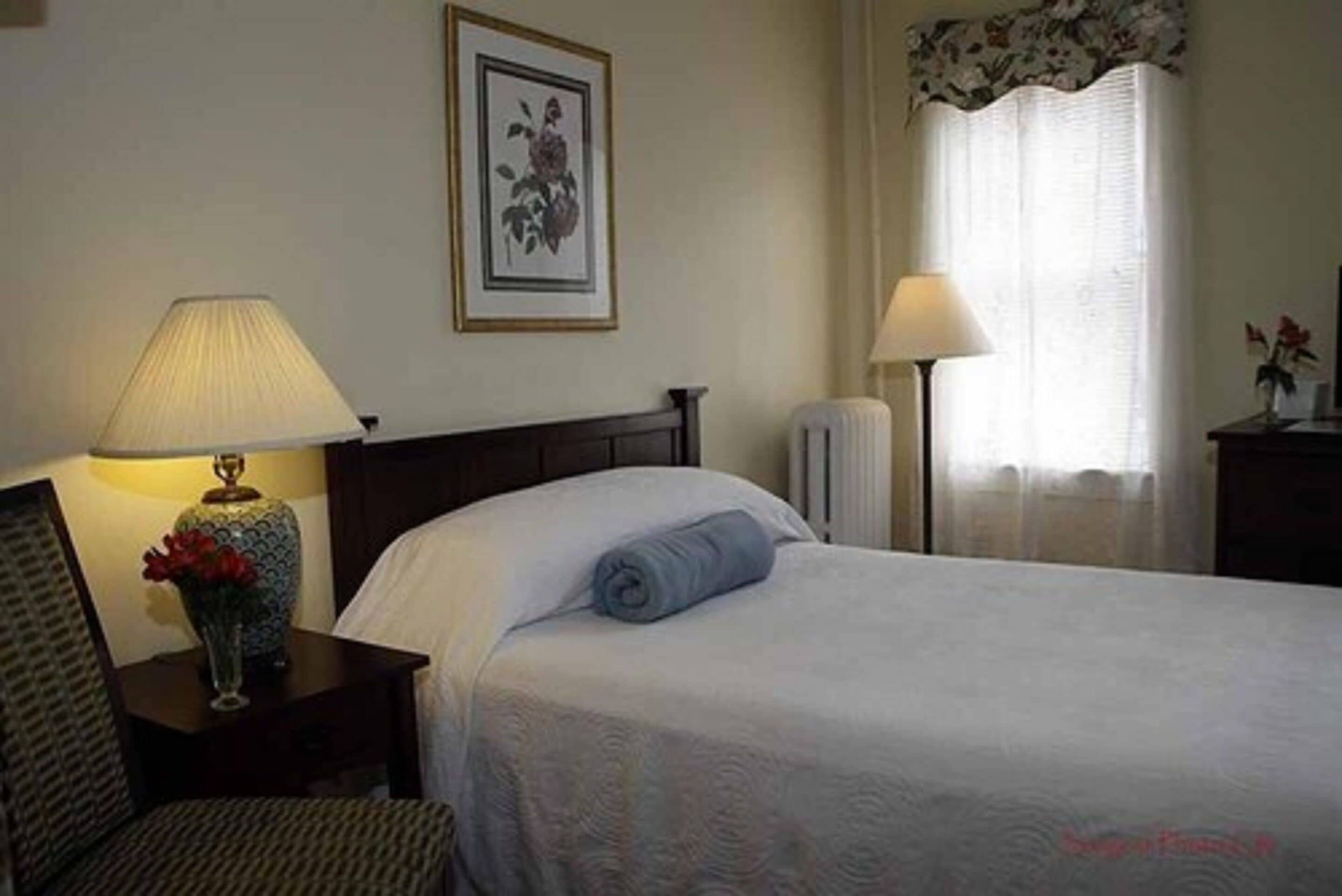 A hotel room with a bed, a nightstand with a lamp, a framed picture on the wall, and a window with curtains.