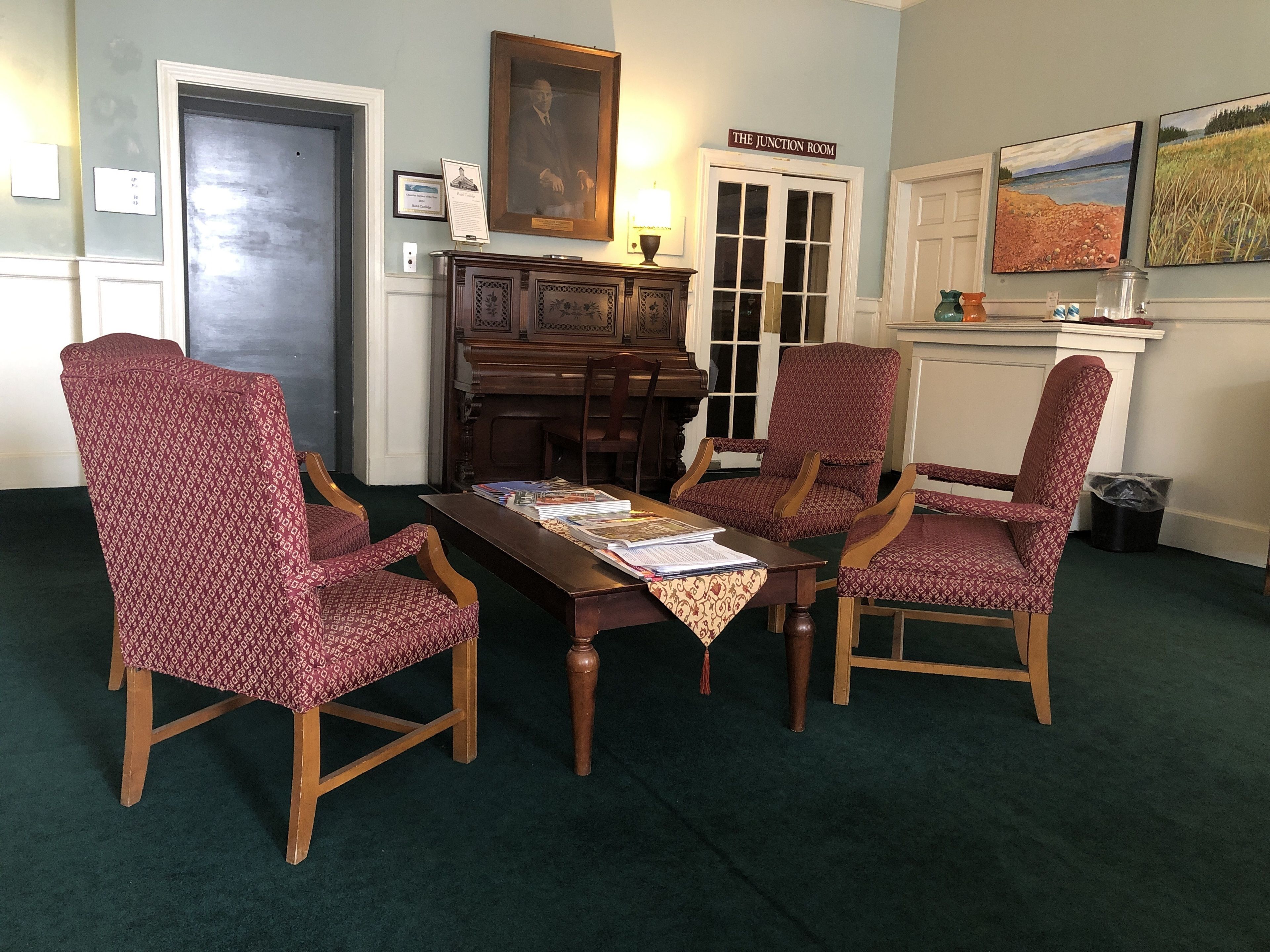 A room with a piano, three patterned chairs, a table with papers, and two paintings on the wall.