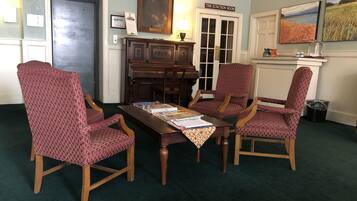 A room with a piano, three patterned chairs, a table with papers, and two paintings on the wall.
