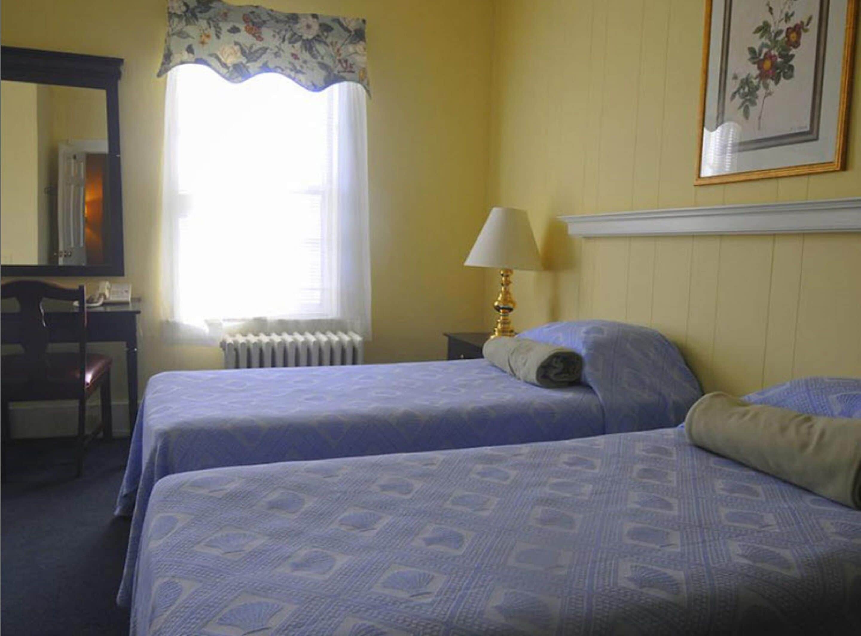 Une chambre d’hôtel avec deux lits, un bureau, une chaise, un radiateur, une lampe et un tableau encadré au mur.
