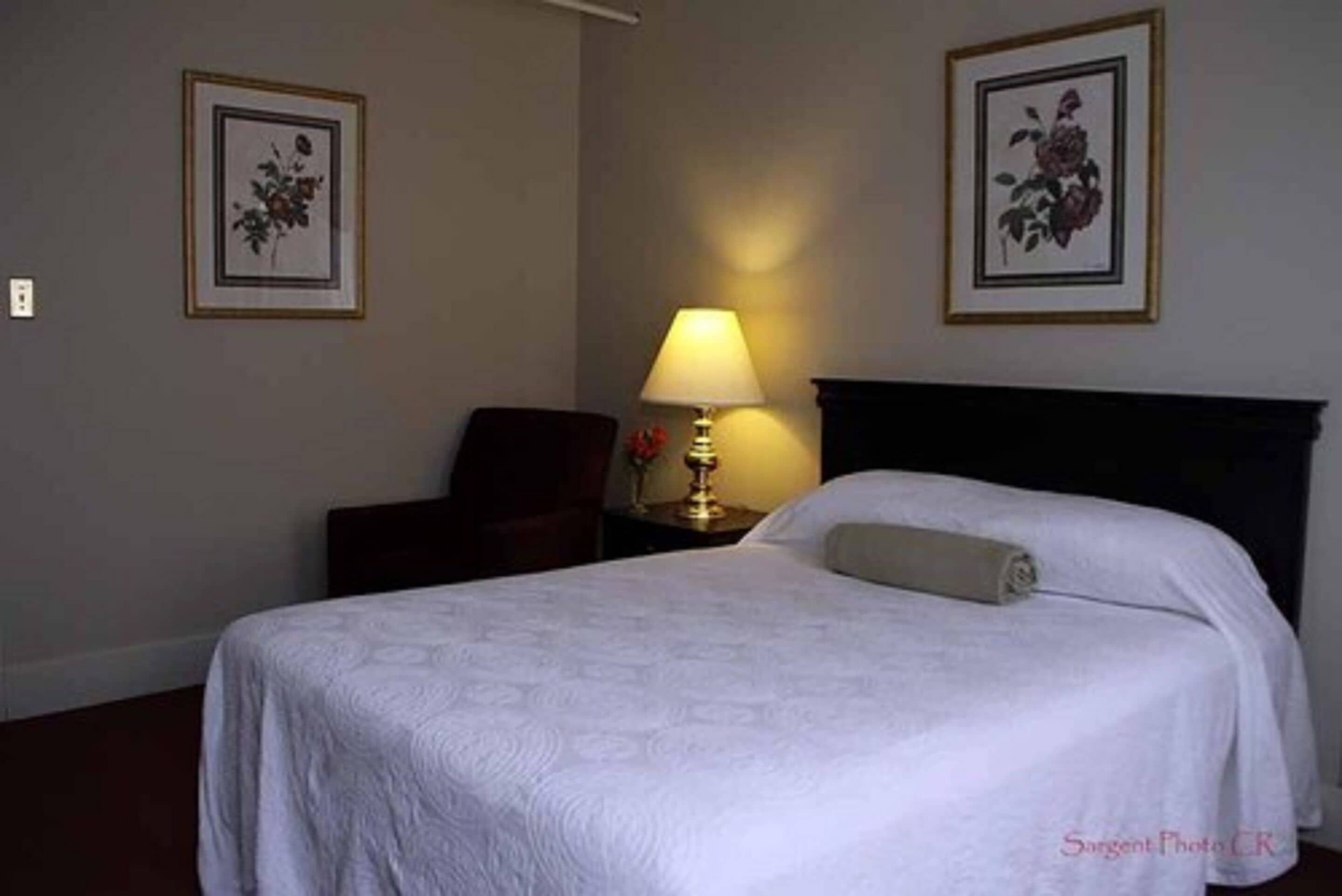 A neatly made bed with a white bedspread, a bedside lamp, and two framed floral pictures on the wall.
