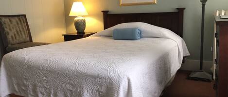 A neatly made bed with a white bedspread, a blue pillow, and a wooden headboard. There is a nightstand with a lamp and a framed botanical print on the wall.