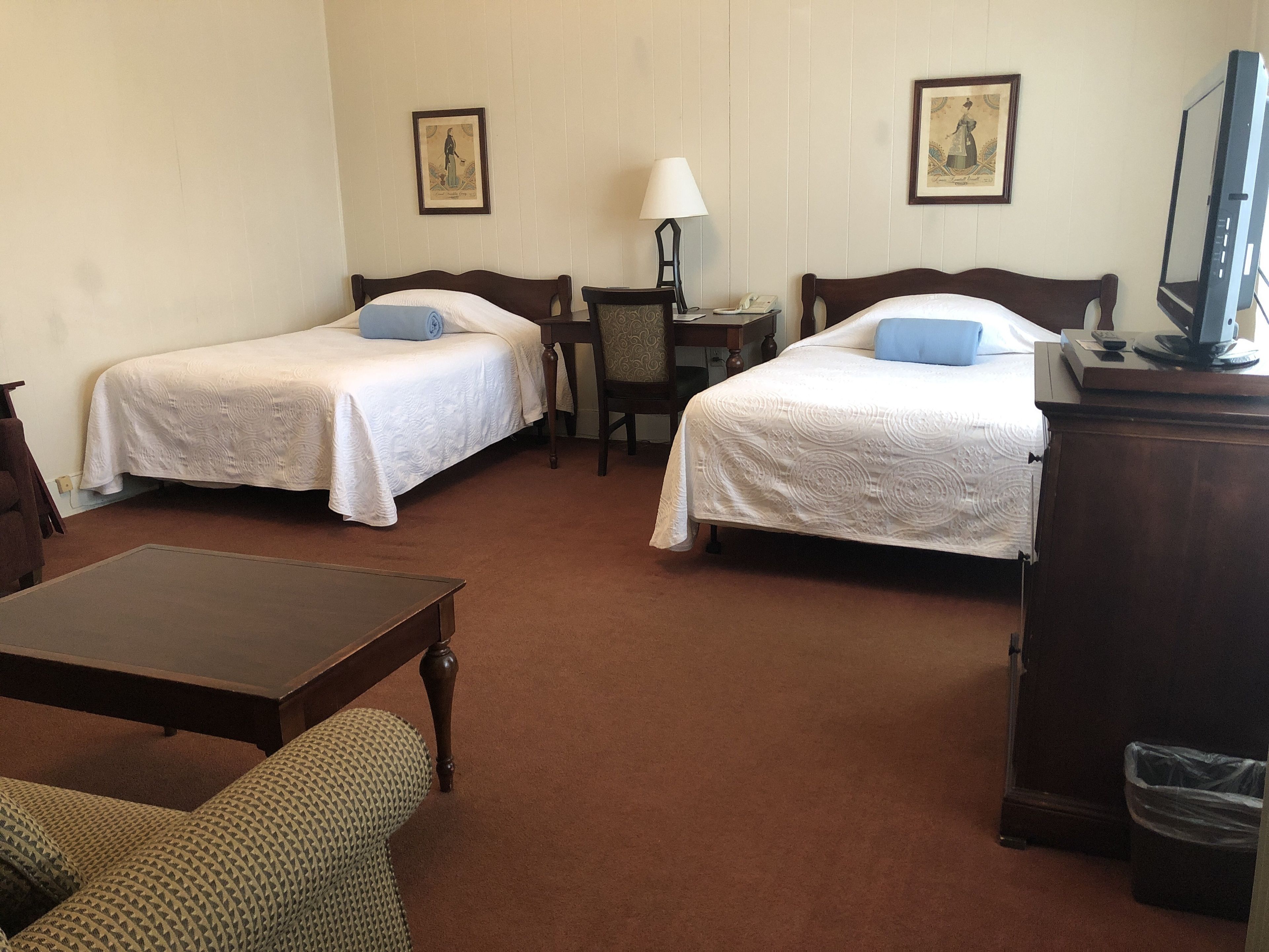 A hotel room with two single beds, a small table, a chair, a TV, and a lamp.