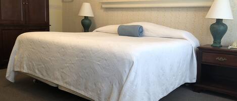 Una cama bien hecha con cubrecama y almohadas blancas, flanqueada por dos mesitas de noche con lámparas, frente a una pared cubierta de papel tapiz.