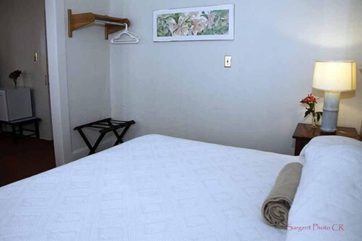 Una cama bien hecha con una toalla enrollada, una mesita de noche con una lámpara, un cuadro en la pared y una silla.