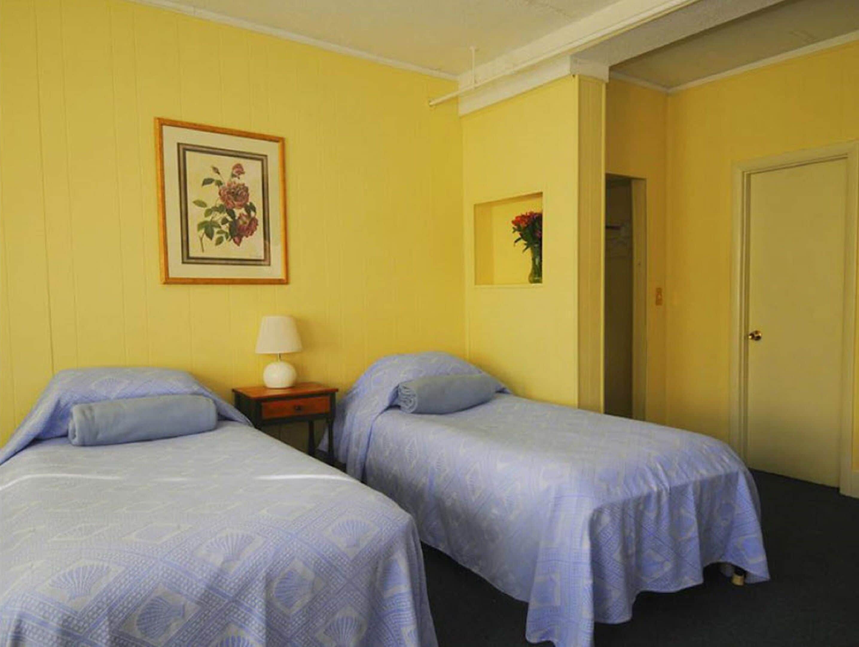 Una habitación con dos camas, una mesita de noche con lámpara, un cuadro de flores enmarcado en la pared y una puerta a otra estancia.
