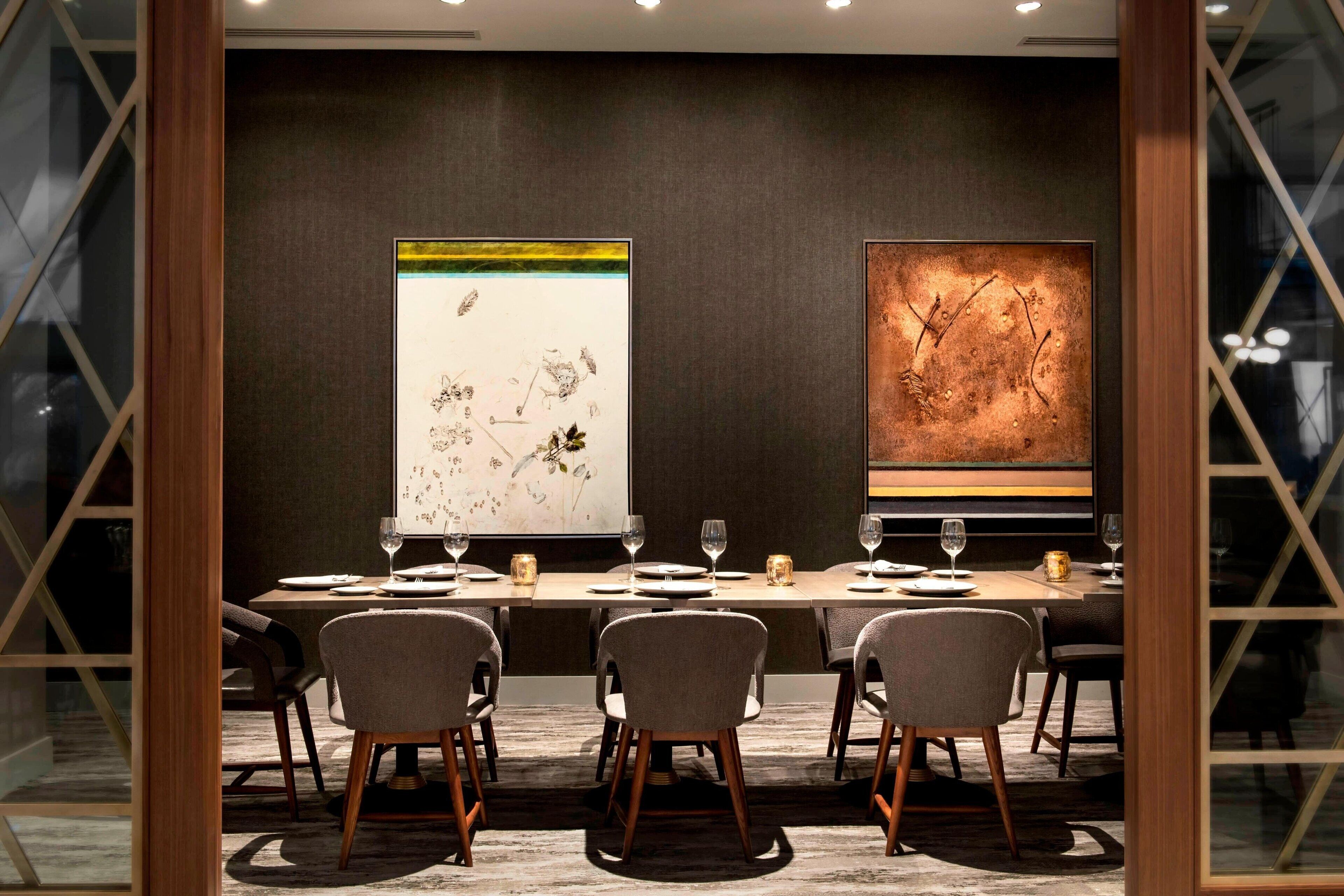 A dining area with a long table set for six, flanked by two large abstract paintings, and a modern interior design with a dark wall and wooden accents.