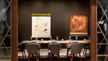 A dining area with a long table set for six, flanked by two large abstract paintings, and a modern interior design with a dark wall and wooden accents.