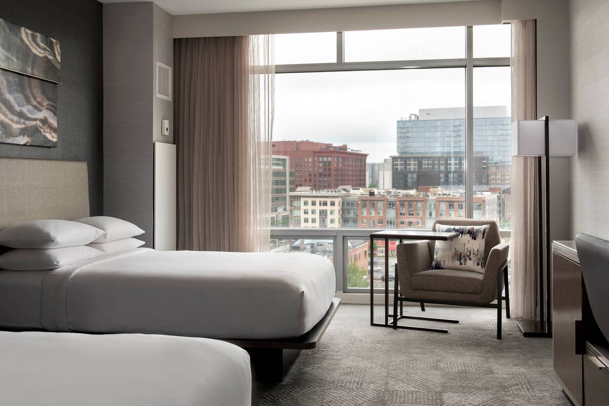 Une chambre d’hôtel avec un grand lit, une chaise, une table et une vue sur la ville grâce à de grandes fenêtres.