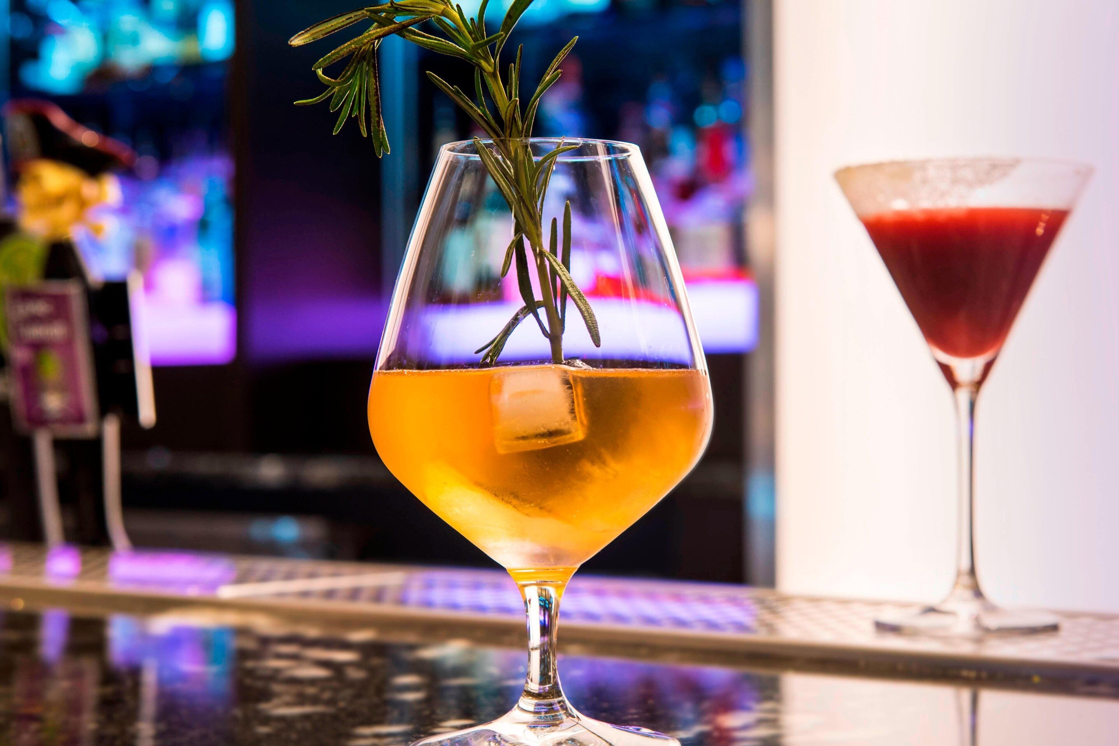 Um copo de coquetel com um ramo de alecrim e um cubo de gelo, apoiado em um balcão de bar com um fundo desfocado de um bar e uma tela de televisão.