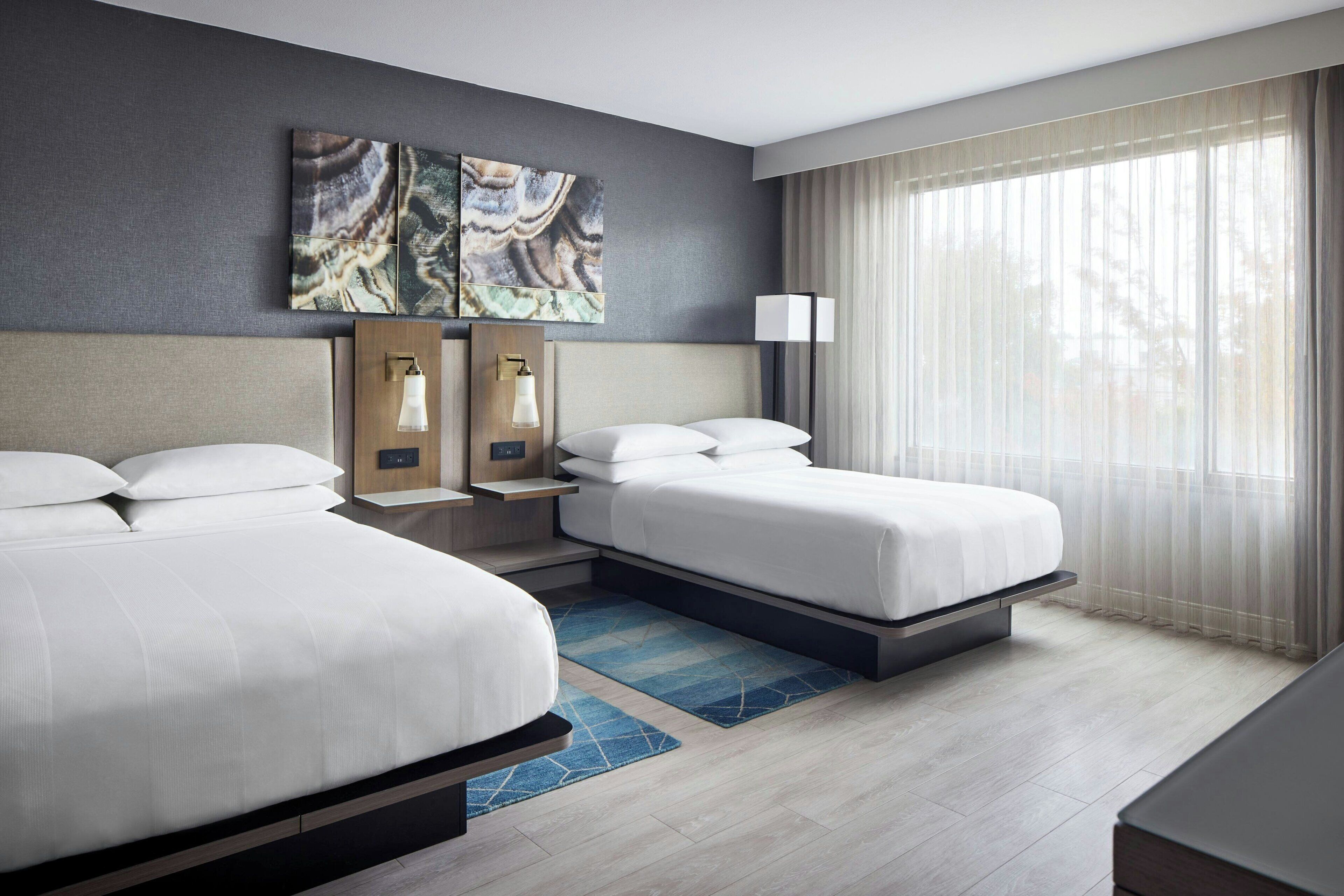 Habitación de hotel con dos camas, un ventanal con cortinas translúcidas, una obra de arte colgada en la pared y una mesita de noche con lámparas.