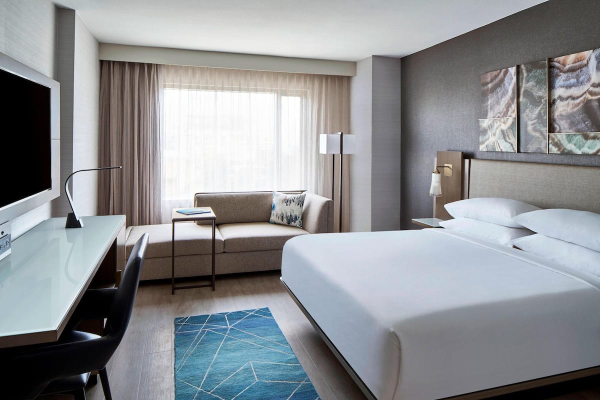 Une chambre d’hôtel avec un grand lit, un bureau avec une chaise, un canapé, un téléviseur à écran plat et une grande fenêtre avec des rideaux.