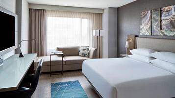 A hotel room with a large bed, a desk with a chair, a sofa, a flat-screen TV, and a large window with curtains.