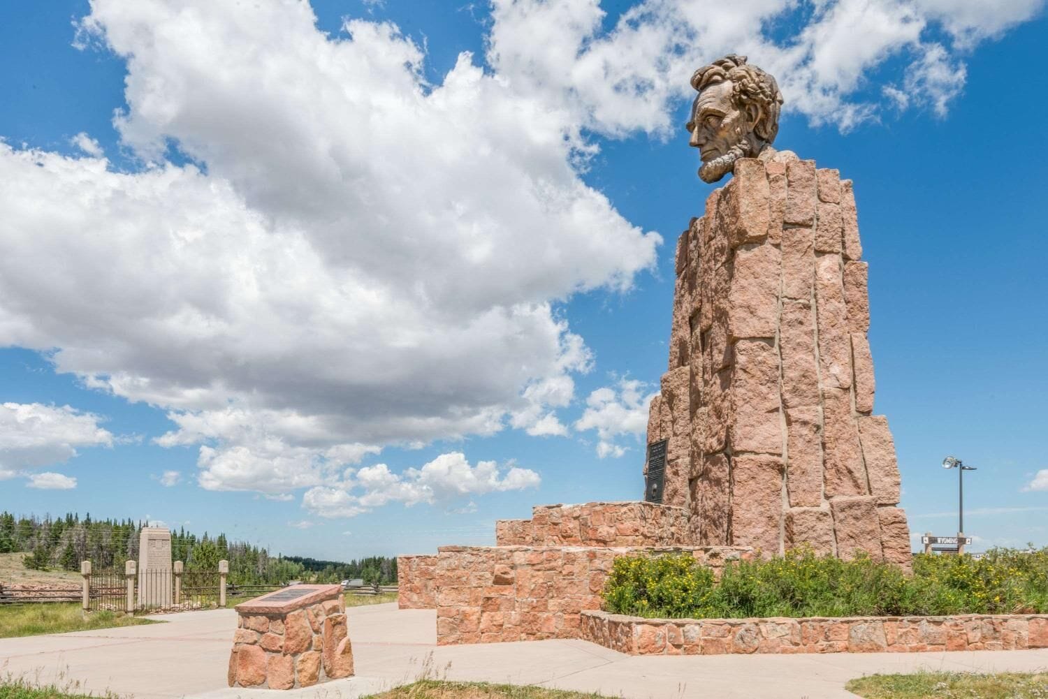 A large stone statue of a historical figure on a pedestal, surrounded by a landscaped area with a clear sky and clouds.