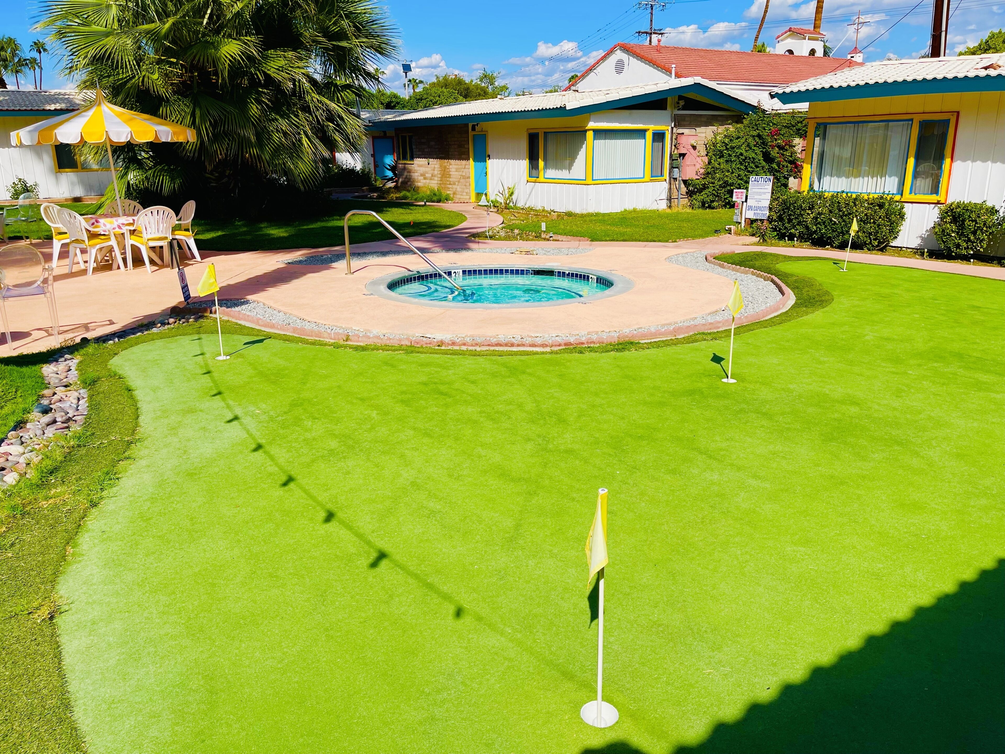 Een goed onderhouden zwembadgedeelte met een putting green, omringd door een huis met een parasol en ligstoelen bij het zwembad.