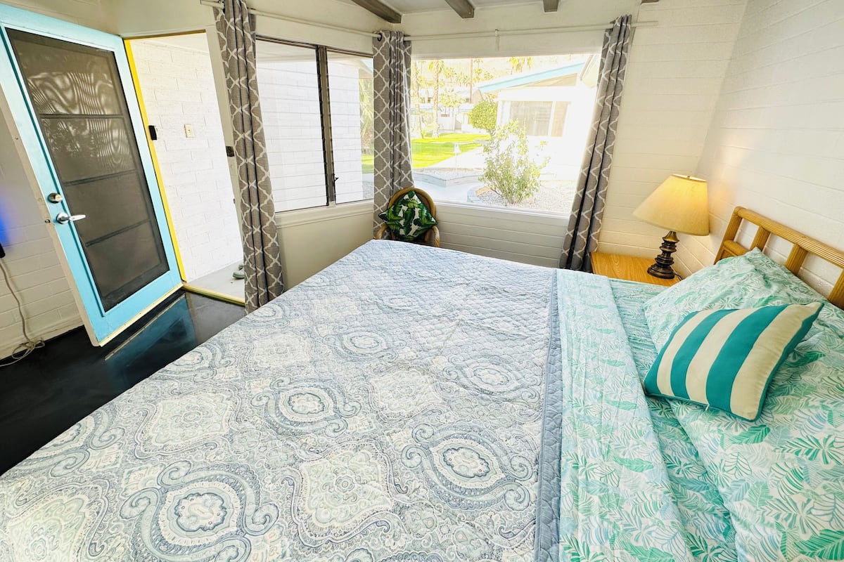 A bedroom with a bed, a window with curtains, a door, and a nightstand with a lamp.