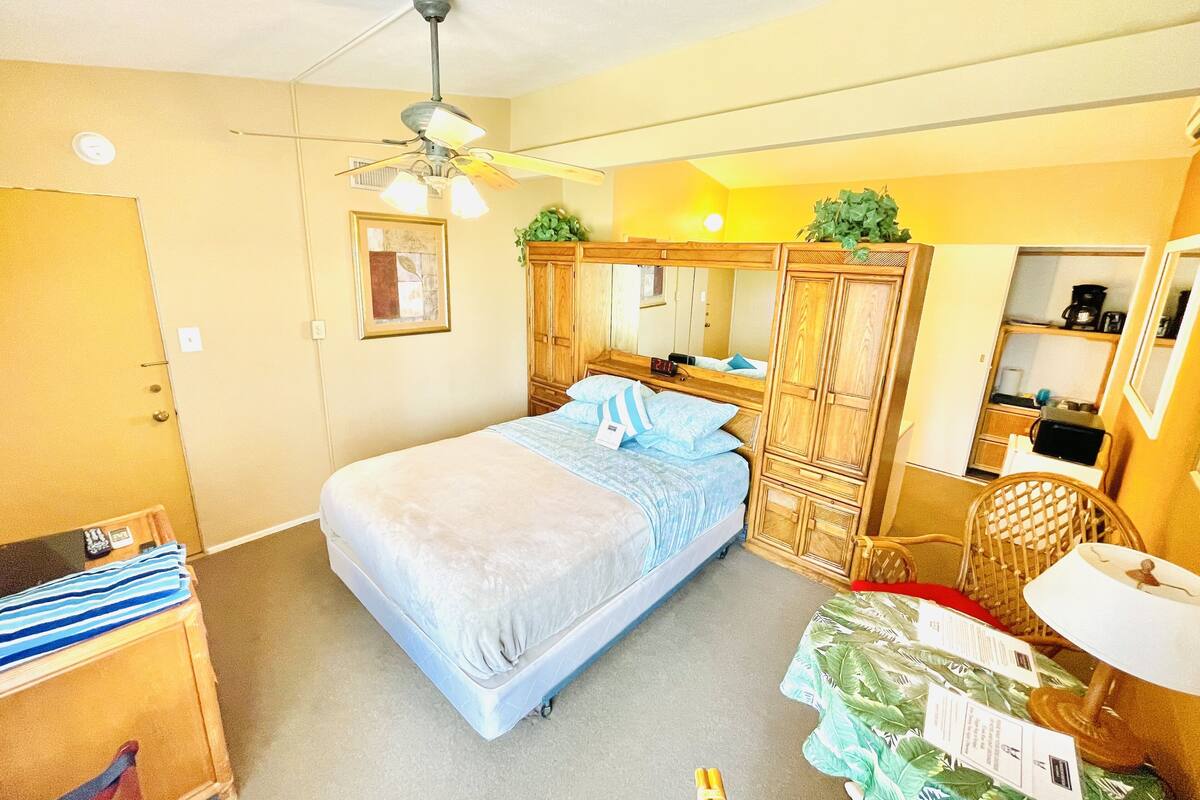 A compact bedroom with a single bed, a ceiling fan, a wooden wardrobe, and a small kitchenette area.