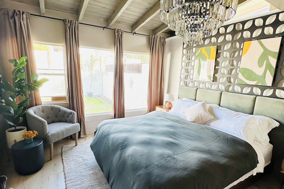 A bedroom with a large bed, a chair, a potted plant, and a chandelier.