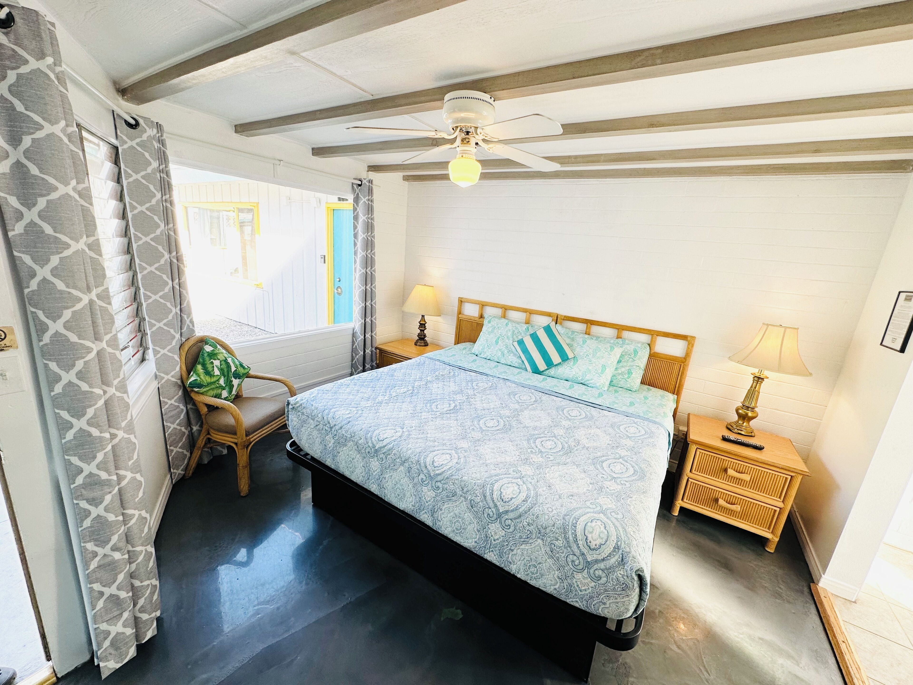 Une chambre à coucher comprenant un lit, une chaise, une table de chevet, un ventilateur de plafond et une fenêtre avec des rideaux.
