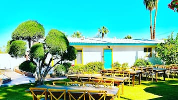A sunny outdoor dining area with neatly arranged wooden tables and chairs, surrounded by well-maintained greenery and palm trees.