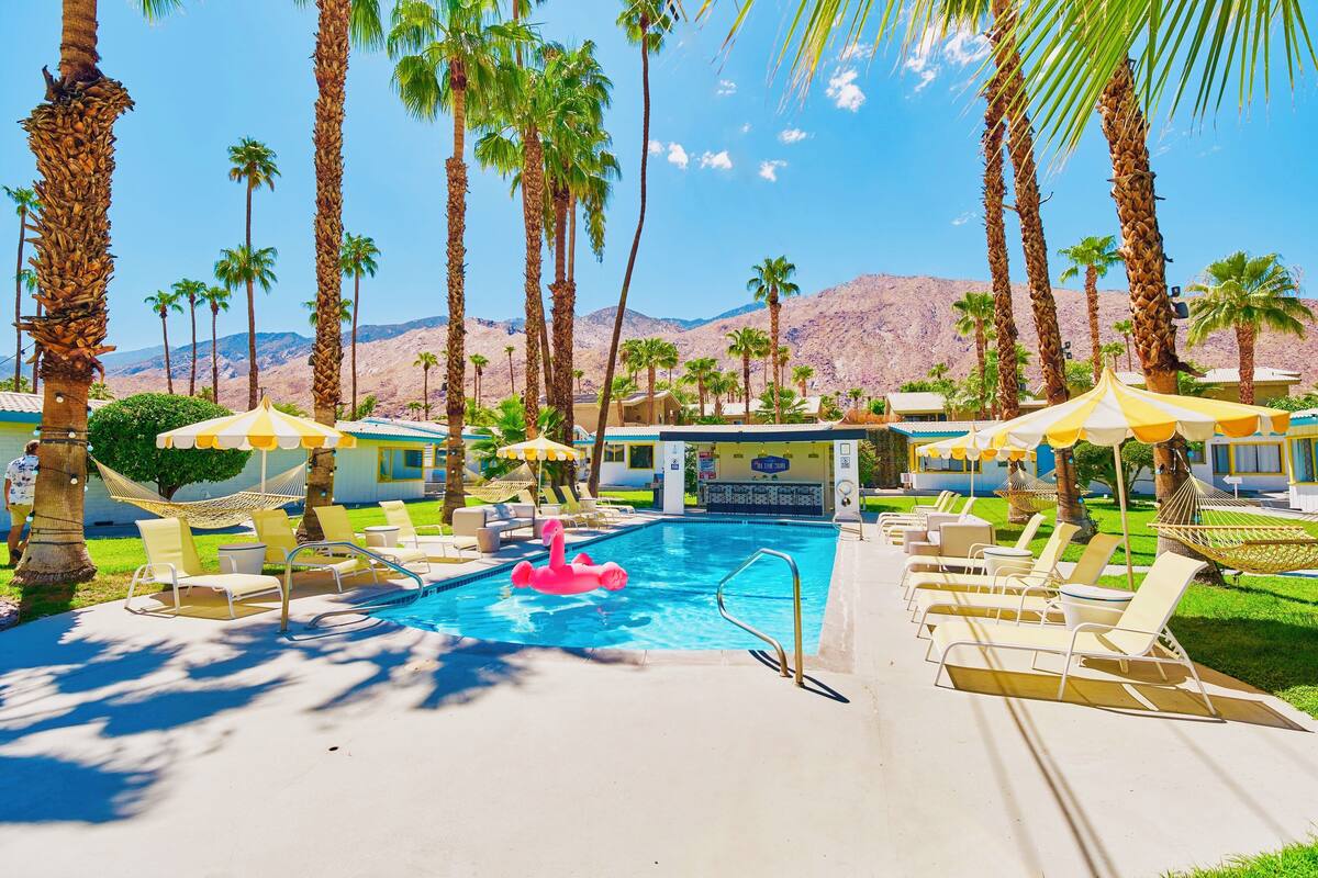 A pool area with lounge chairs and umbrellas, surrounded by palm trees and a mountainous backdrop.