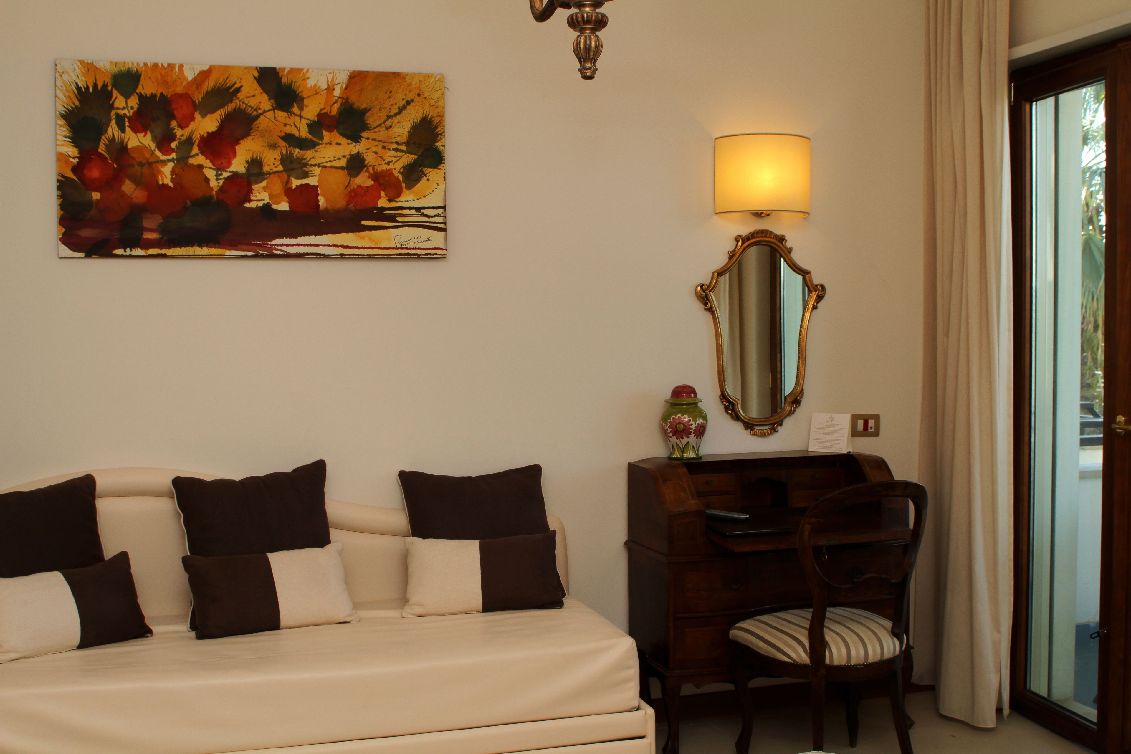A hotel room with a bed, a desk, a chair, a lamp, a mirror, and a painting on the wall.