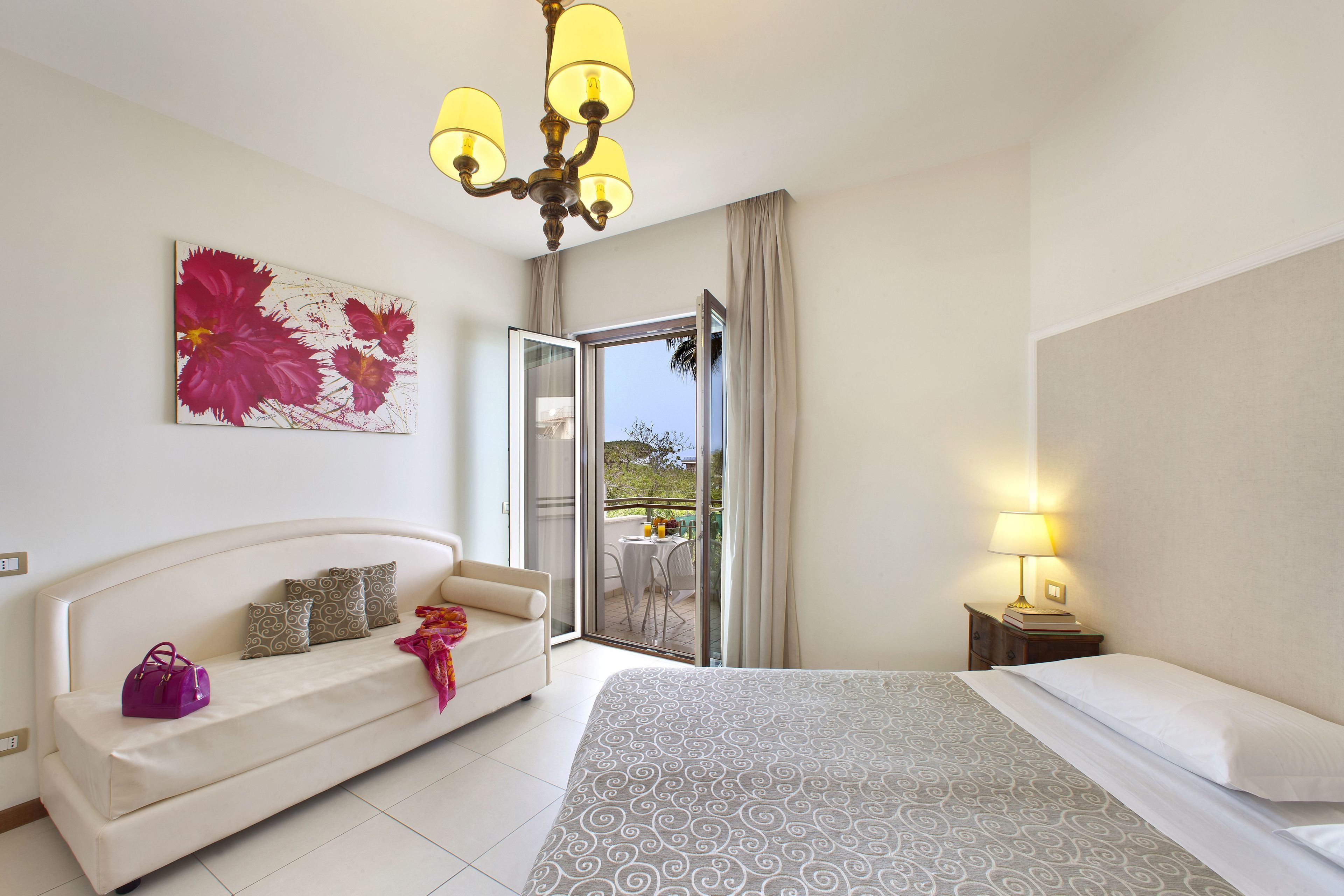 A hotel room with a bed, a sofa, a chandelier, a painting, and a balcony with a table and chairs.