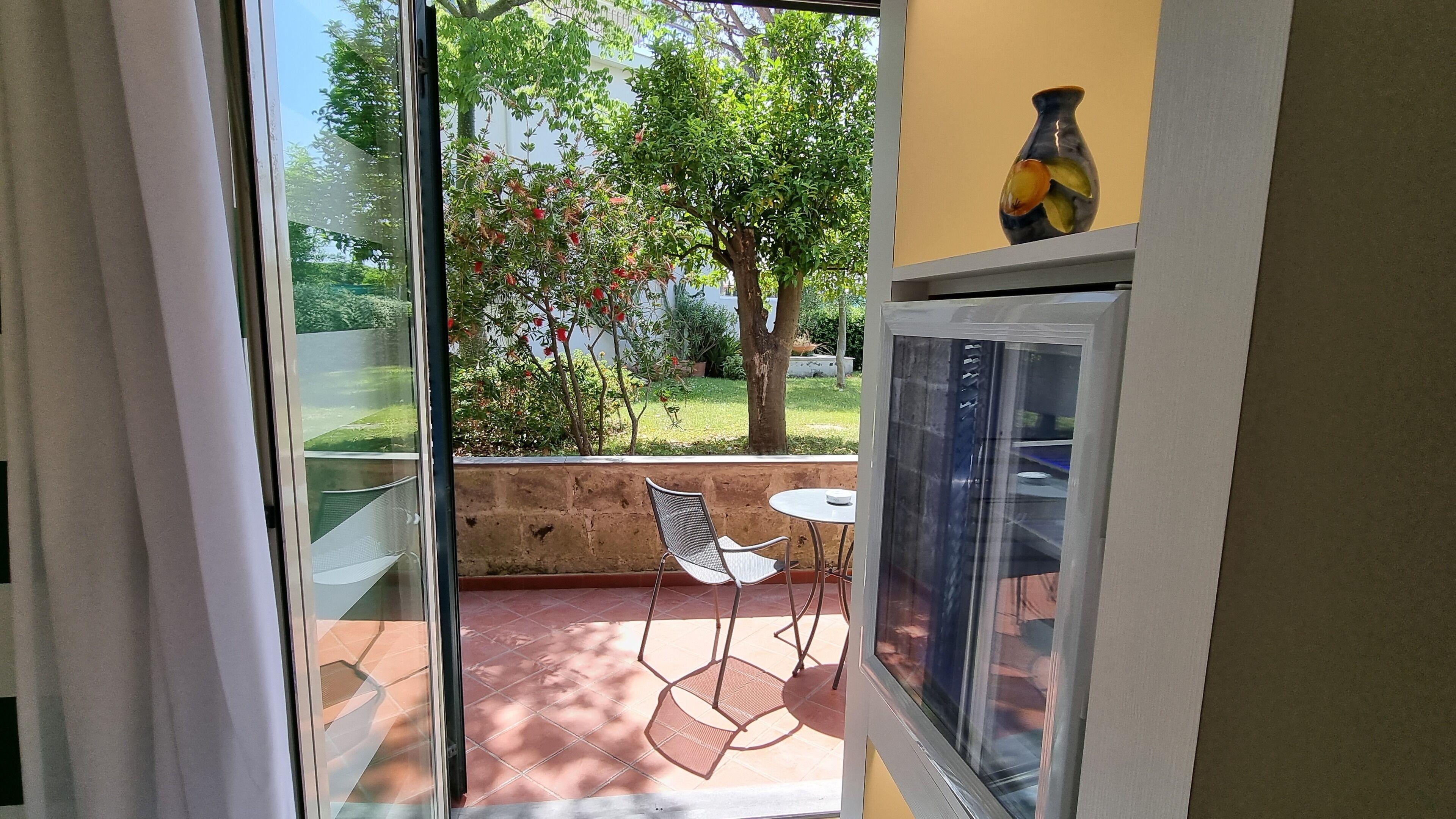 Une porte vitrée donne sur un patio extérieur équipé d’une table et de chaises, ainsi qu’un vase posé sur une étagère et un arbre aux fleurs rouges.
