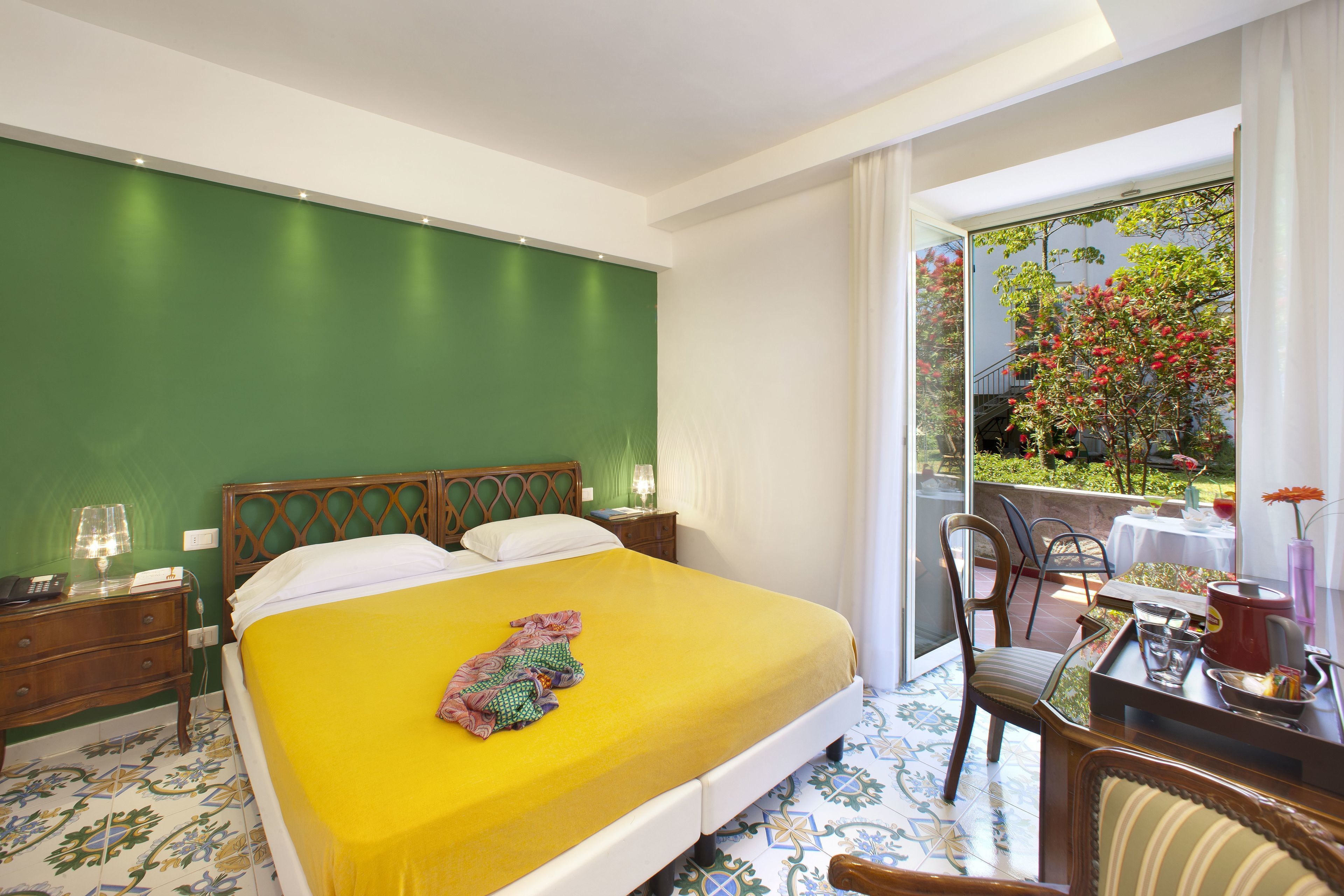 A hotel room with a bed, a desk with a chair, a small table, and a balcony with a view of a garden.