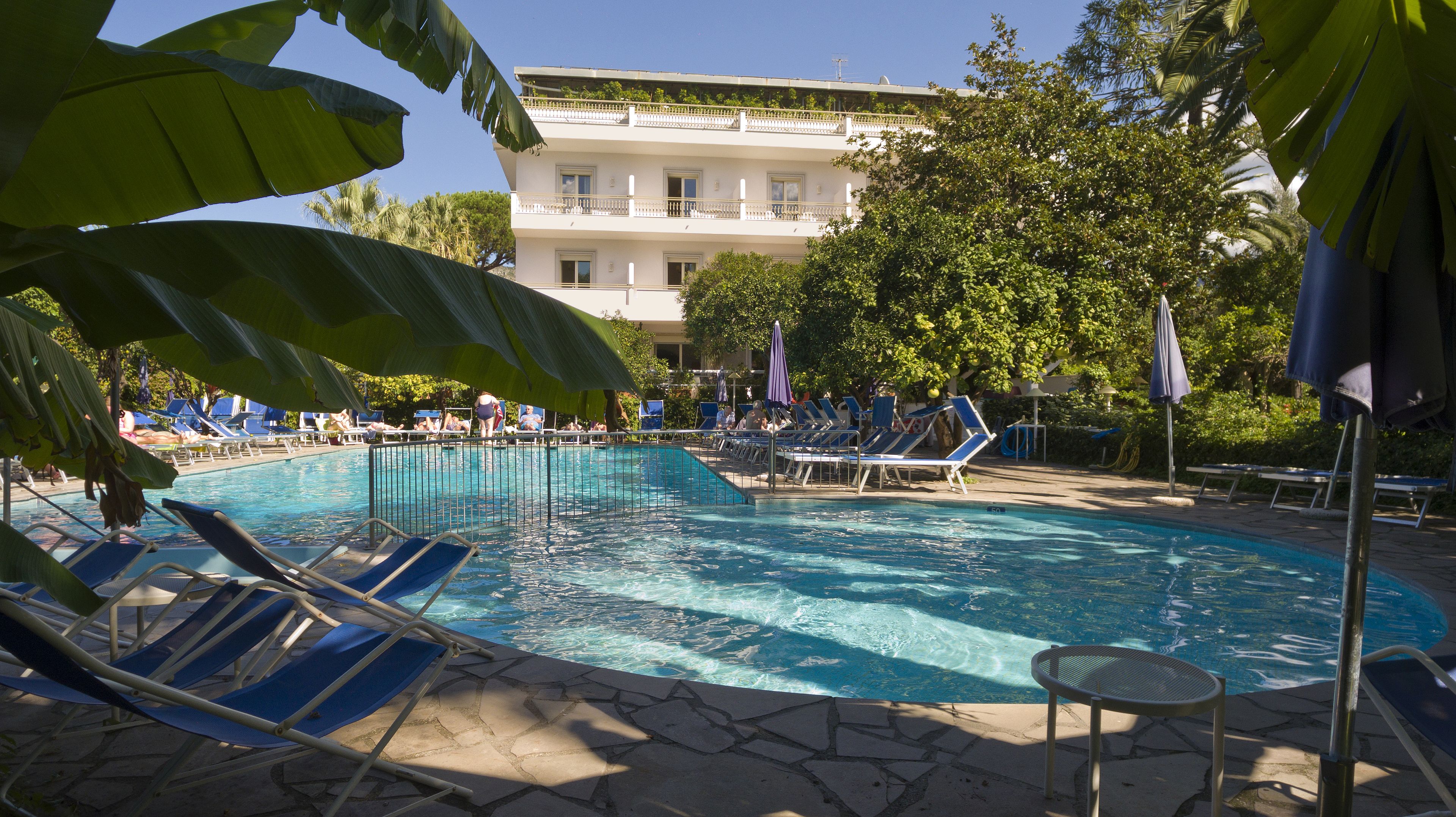 A swimming pool with lounge chairs and umbrellas, surrounded by trees and a building in the background.