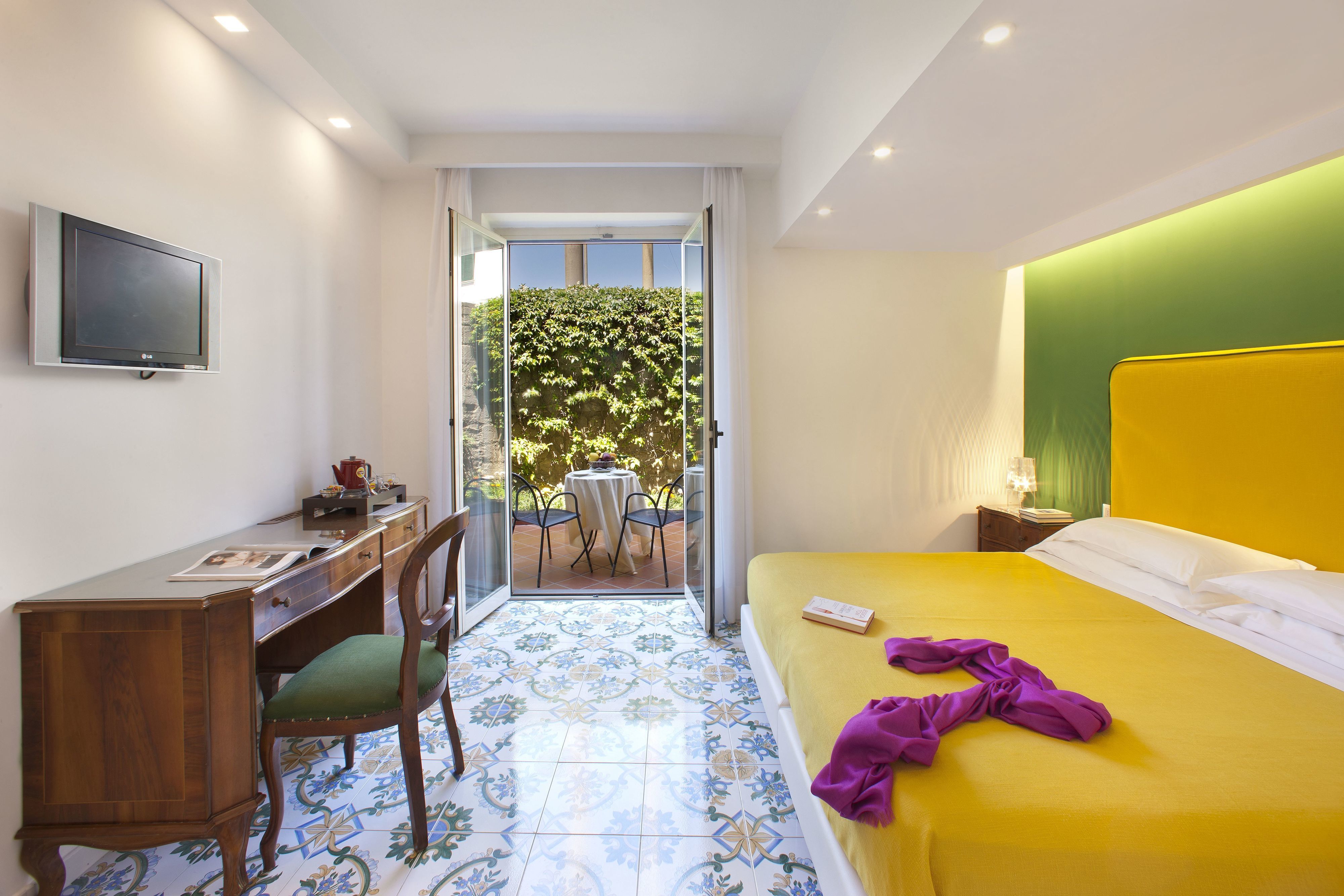 A hotel room with a bed, a desk, a chair, a TV, and a balcony with a table and chairs.