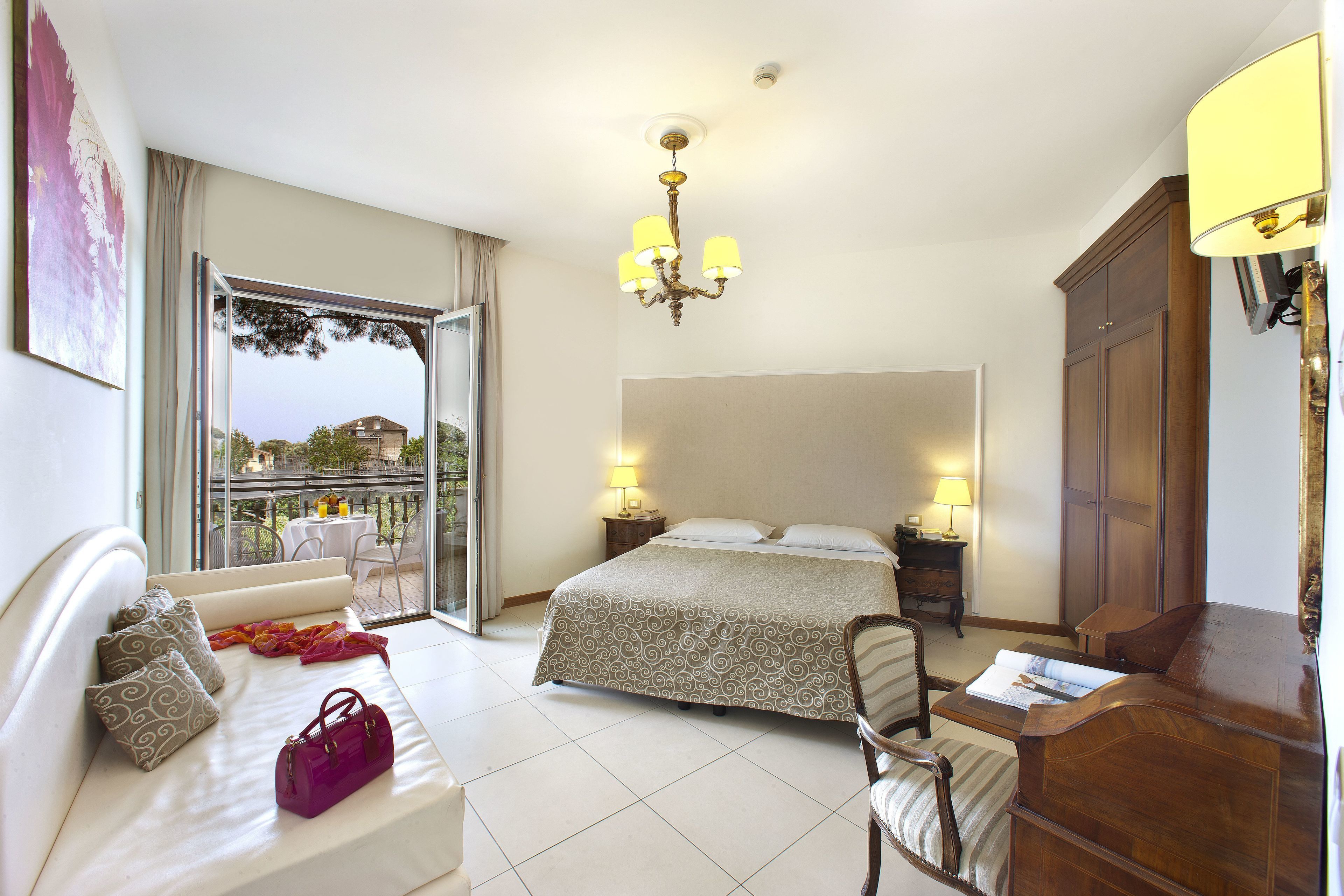 A hotel room with a bed, a sofa, a desk, a chair, a chandelier, and a view of the outside.